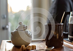 Steamy cup of coffee with sugar cubes and coffee pot in kitchen with morning sunshine