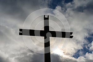 Steel cross against cloudy sky with the sun breaking through and patches of blue