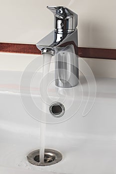 Steel faucet in the bathroom. Stream of water falling in ceramic sink. Modern metal mixer with white wash basin. Open tap.