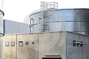 Steel industrial silos for liquids and solids standing in a factory next to a concrete building.