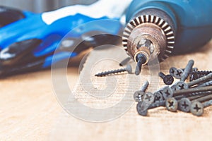 A steel screw is placed on an electric drill near scattered screws on a wooden board background. The concept of tools and repair