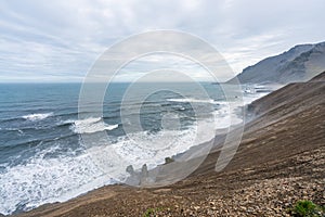 Steep cliffside with volcanic beach and waves crashing to shore. Mountains in the background. photo