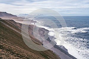 Steep cliffside with volcanic beach and waves crashing to shore. Road and mountains with snow in the background.