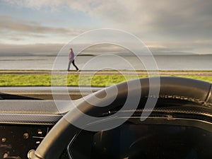 Steering wheel in focus. Nature scene with ocean, mountain and a man walking by out of focus. Travel by car concept