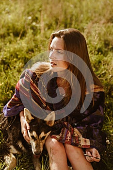 On the steppe grass sits a long-haired blonde beauty with no makeup and hugging the dog looking into the distance