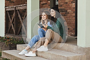 On the steps near the house, two beautiful girls-sisters who have come from the university are sitting, use a tablet and laugh.