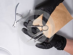 Sterilized manicure items in kraft paper packaging. Hands in black surgical protective gloves hold manicure devices.