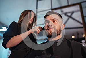 Stern bearded Man portrait while undercut hairstyle hairdressing in Hair Salon by young hairdresser female. Modern low light black