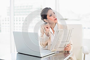 Stern businesswoman holding newspaper while working on laptop lo