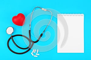 Stethoscope with blank notepad, red heart and white capsules on a blue background.