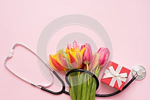 Stethoscope with bouquet of tulips and little red gift box on light pink background with a copy space