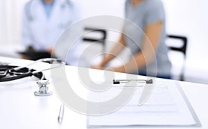 Stethoscope, clipboard with medical form lying on hospital reception desk with laptop computer and busy doctor and