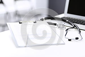 Stethoscope, clipboard with medical form lying on hospital reception desk with laptop computer and busy doctor and