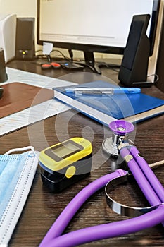 Stethoscope, face mask and pulse oximeter on the background of a notebook and a computer monitor - a doctor`s workplace