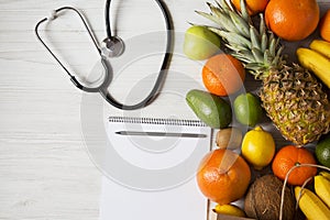 Stethoscope health diet. Paper bag of different fruits with notepad on white wooden background.
