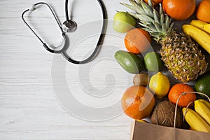 Stethoscope health diet. Paper bag of different fruits on white wooden background. Flat lay.