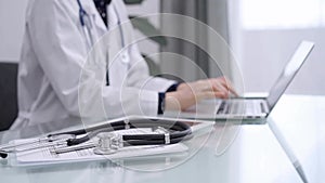 Stethoscope and medical clipboard are lying on the glass table while doctor woman is working with laptop computer on the