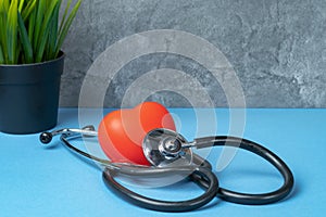 Stethoscope and rubber red heart on a blue table with a plant against a gray marble wall.