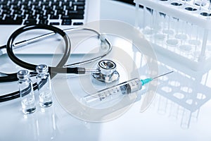 Stethoscope, syringe, vials, and laptop on a white table.