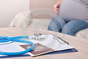 Stethoscope and ultrasound scan on the table, pregnant woman sitting on the sofa on the background