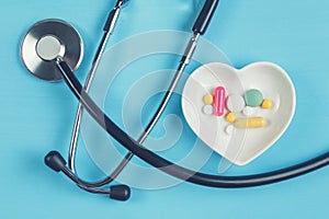 Stethoscope and various pills in a saucer in shape of heart on a blue wooden background