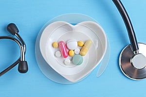 Stethoscope and various pills in a saucer in shape of heart on a blue wooden background