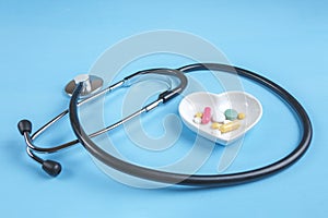 Stethoscope and various pills in a saucer in shape of heart on a blue wooden background