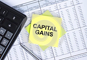 Sticker with text Capital Gains lying on the financial tables. Calculator with metal pen