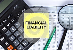 Sticker with text Financial Liability lying on the calculator. Magnifying glass with green pen on financial documents