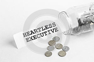 Sticking out of a jar of coins a piece of paper with a text Heartless Executive on a white background