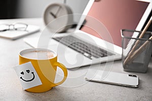 Sticky note with funny face attached to cup of coffee on office table