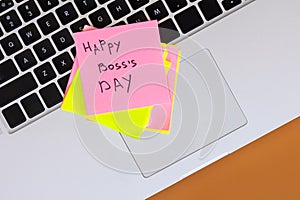 Sticky note with phrase Happy Boss Day attached to laptop on brown table.