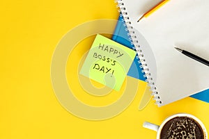 Sticky note with phrase Happy Boss Day attached to notebook on yellow table.