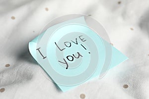 Sticky note with phrase I love you! on bed, closeup