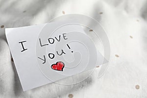 Sticky note with phrase I love you! on bed, closeup