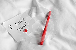 Sticky note with phrase I love you! and red felt pen on bed, space for text