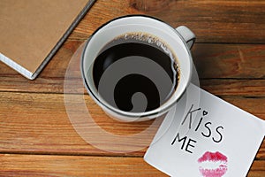 Sticky note with phrase Kiss Me, lipstick mark and cup of coffee on wooden table, above view