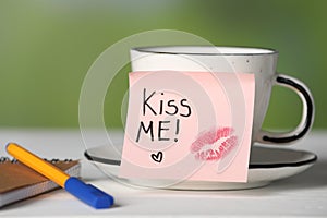 Sticky note with phrase Kiss Me, lipstick mark, cup of drink and notebook on white table against blurred background, closeup