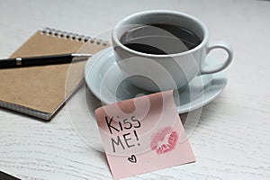 Sticky note with phrase Kiss Me, lipstick mark, cup of drink and notebook on white wooden table