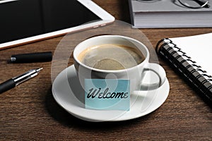 Sticky note with word Welcome attached to cup of coffee on wooden office desk