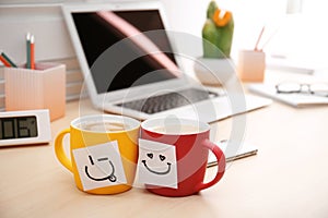 Sticky notes with funny faces attached to cups of coffee on office table.