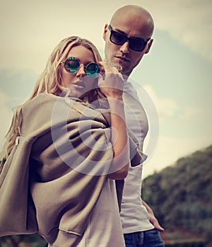 Stilish couple walking in modern dresses and trendy sunglasses on summer background. Beautiful female blond model with hoody on