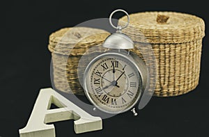 Still Life of Alarm Clock with two wicker boxes and a capital letter A in matt white and corporeal wood
