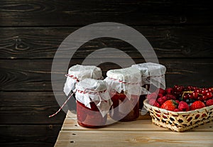 Still life with assorted berries in a basket, glass jars with jams from different berries on wooden table