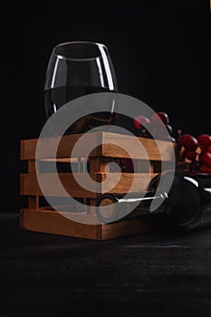Still life of a bottle and a glass of red wine inside a wooden box with grapes, black background