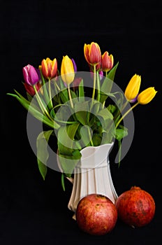 A still life with a bouquet of colourful tulips in a white vase with two pomegranates in front of black background