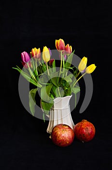 A still life with a bouquet of colourful tulips in a white vase with two pomegranates in front of black background