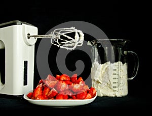 A still life with a bowl of fresh an red strawberries, a food processor, mixer whose wire wisks are full of cream, beside a beaker