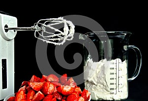 A still life with a bowl of fresh an red strawberries, a food processor, mixer whose wire wisks are full of cream, beside a beaker