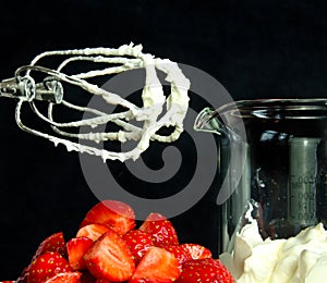 A still life with a bowl of fresh an red strawberries, a food processor, mixer whose wire wisks are full of cream, beside a beaker